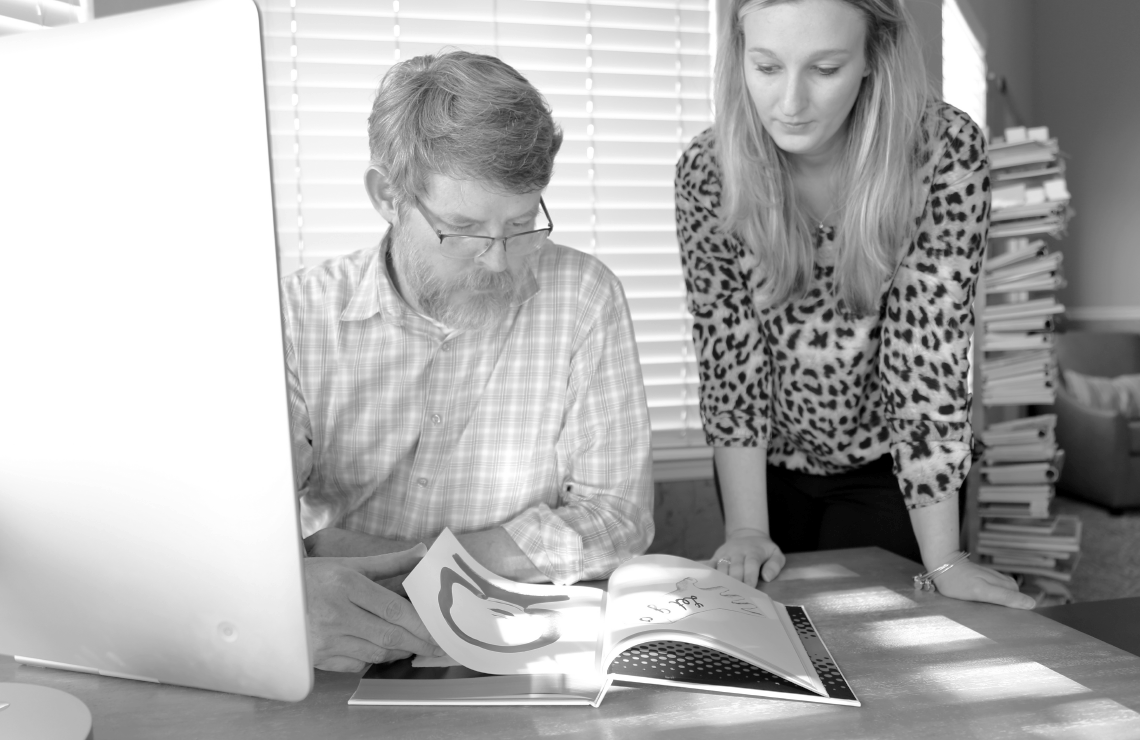 A man sitting and woman standing at a desk, looking at a design book.