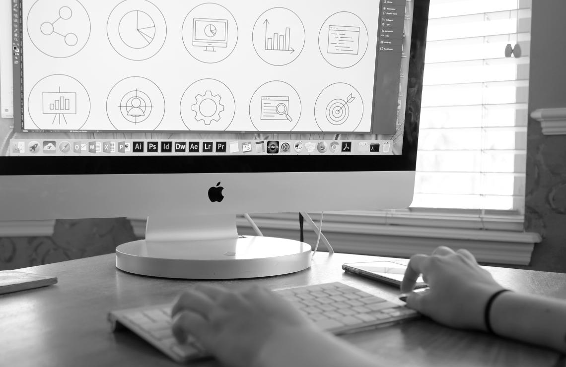 A desktop computer screen showing icon designs in Adobe Illustrator with a designer’s hands on the keyboard and mouse in the foreground.