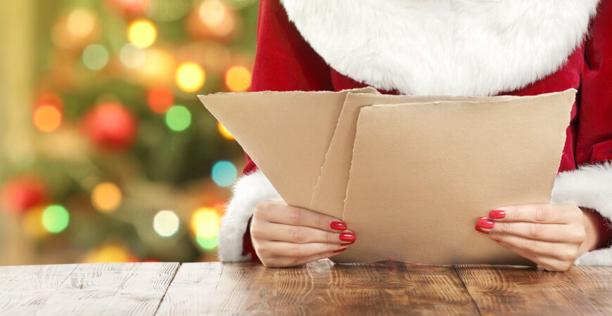 Close up of a woman wearing a Santa outfit with painted red nails sitting at a wood table holding three sheets of brown paper that have torn edges, with a blurred Christmas tree with lights in the background.