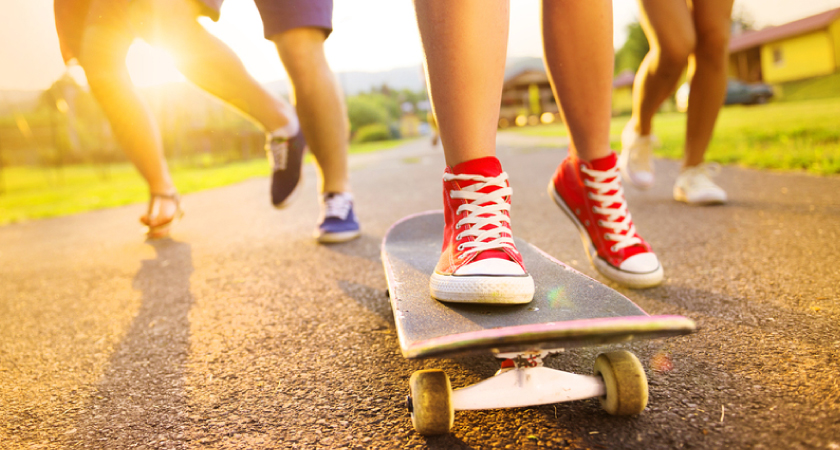 Close up of the feet of three people running along a road at sunset, all wearing sneakers, with a person at the front skateboarding and wearing bright red high-tops.