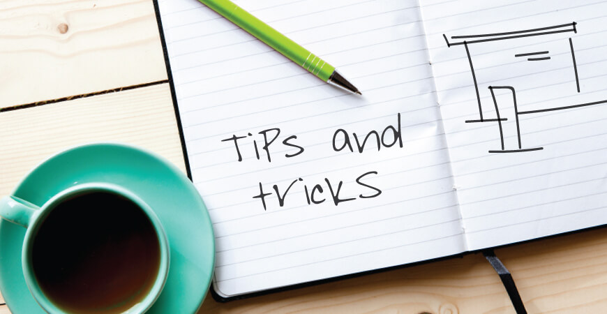 Top view of a teal full cup of coffee next to an open notebook with a green pen and sketch that reads “tips and tricks” and a loose display sketch all sitting on a light wood table.