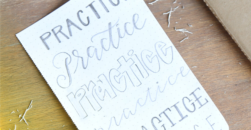 Close up of a dotted piece of paper that has different graphic stylings of the word “Practice” written in pencil with paper shavings around it on a wood desk.