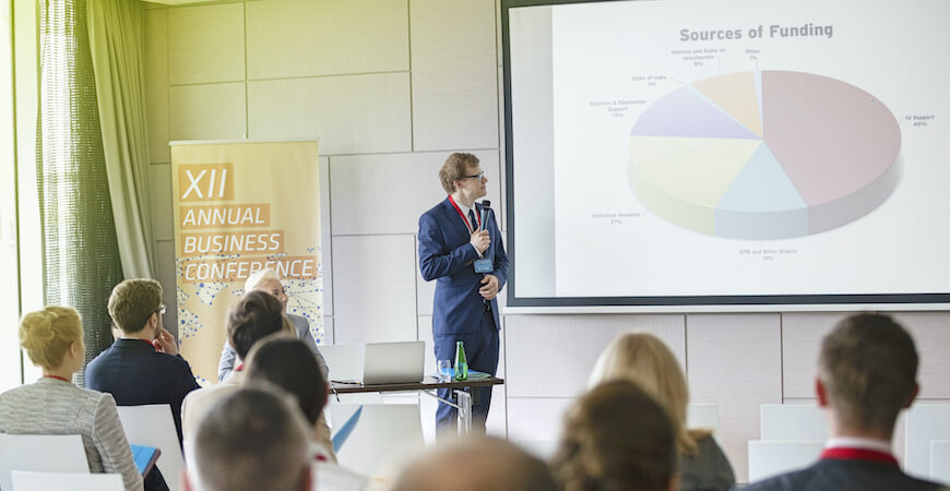 A man standing giving a presentation at a business conference looking at a screen with a pie chart, with an audience in the foreground.