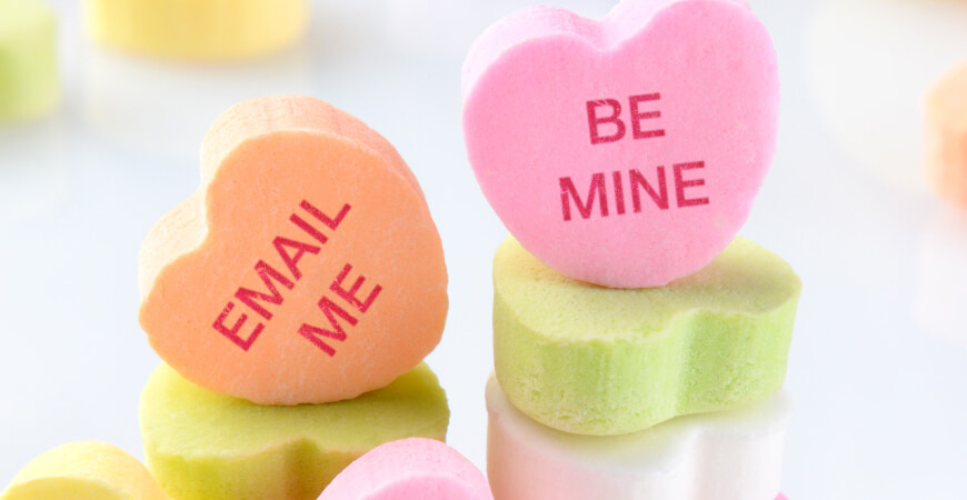 Close up of candy hearts sitting on other candy hearts that read, “Email Me” and “Be Mine” in soft light.